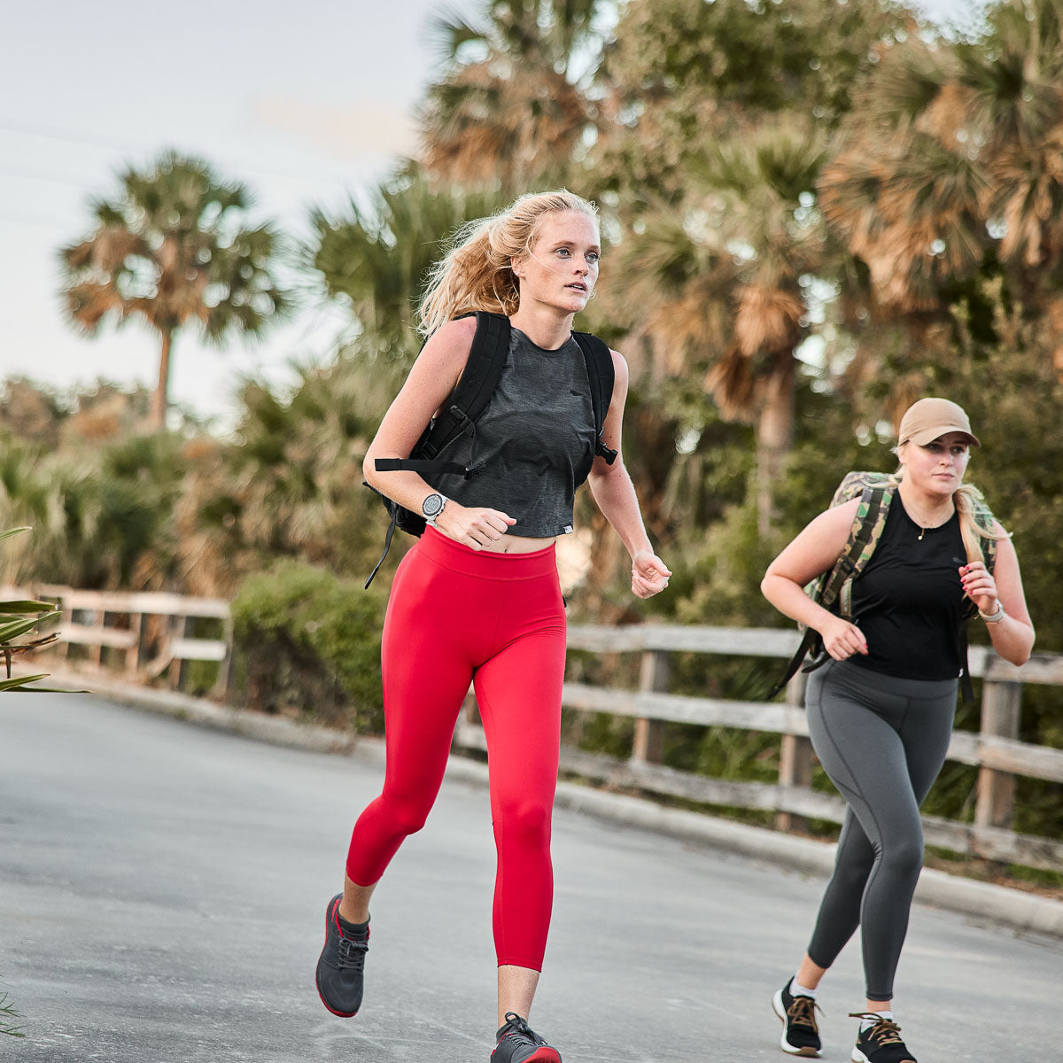 Two women jog beside palm trees, focused, wearing squat-proof Women’s Training Leggings - ToughFlex and backpacks.