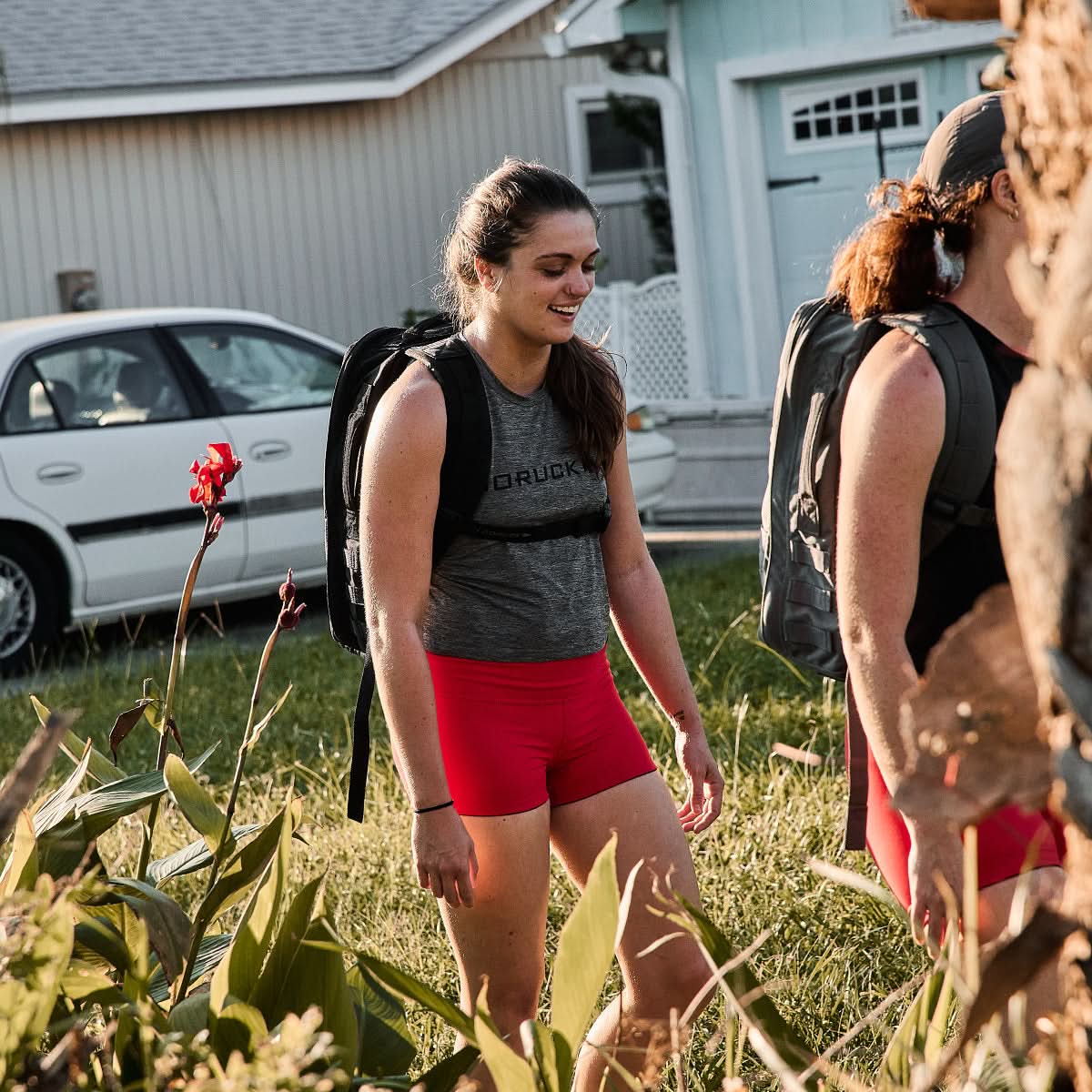 Two individuals are walking outside with backpacks, near a house and car surrounded by green plants and flowers. They're wearing GORUCK Women’s Squat Shorts made with ToughFlex fabric, which provides comfort and mobility on their journey. With stay-put technology, they're prepared for any adventure ahead.