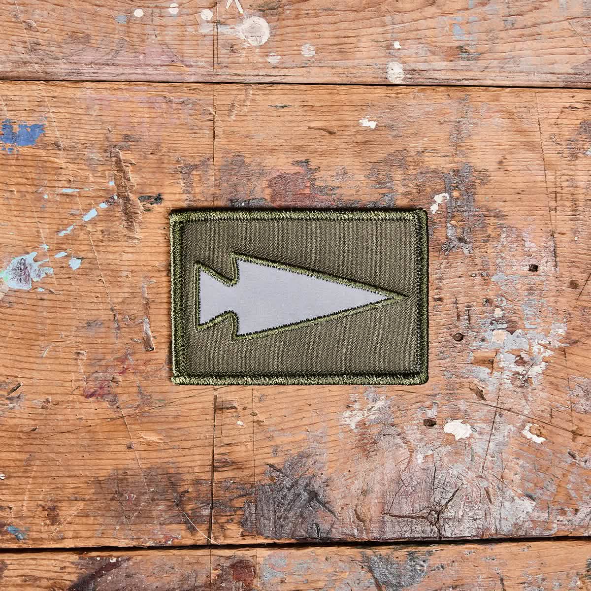 The Patch - Reflective Spearhead by GORUCK, featuring an olive green background with a gray arrowhead design, is displayed on a rustic, weathered wooden surface. The patch is surrounded by paint splatters and scratches on the wood, and it includes a VELCRO backing for easy attachment.