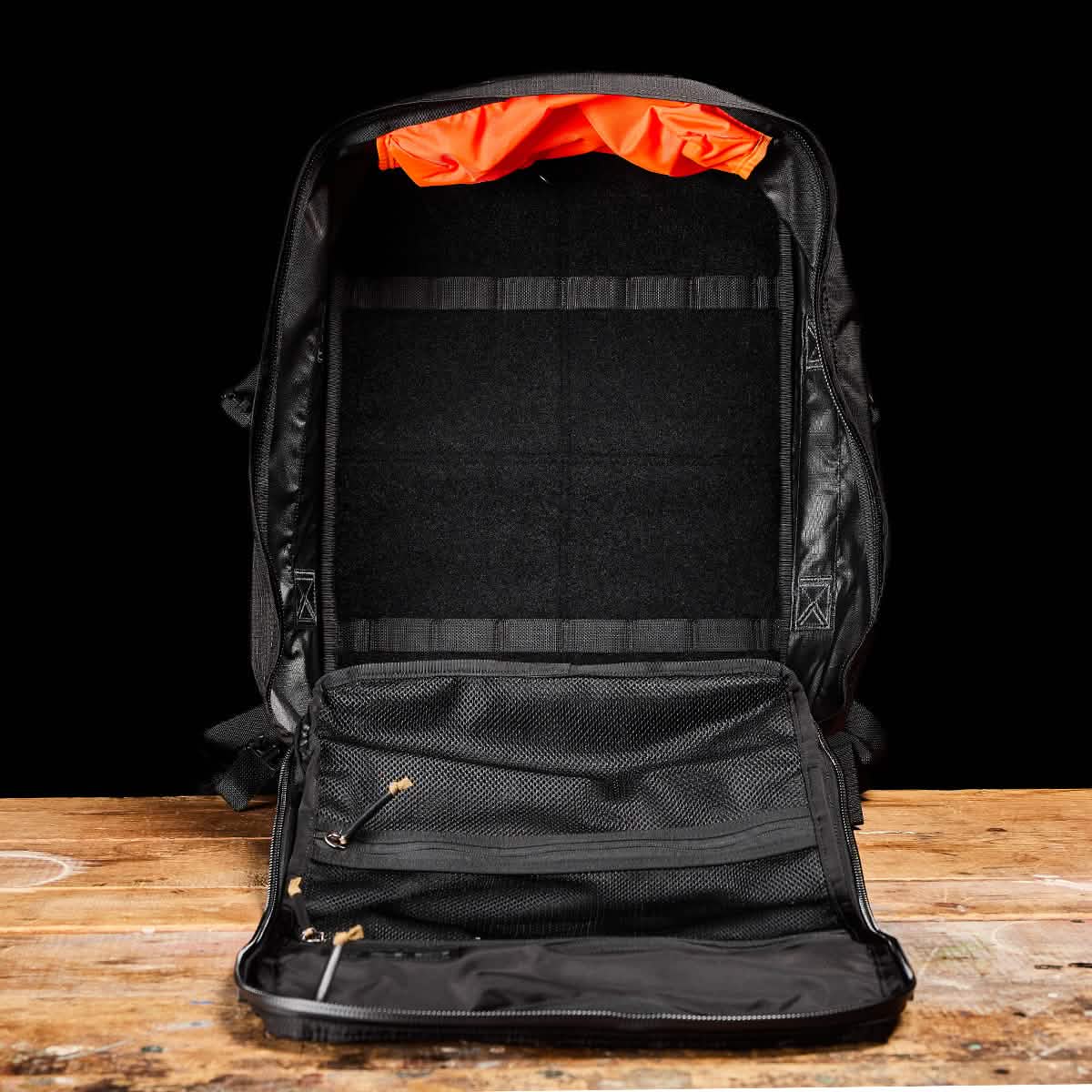 The unzipped black GR3 - Ripstop ROBIC® backpack, designed by GORUCK and made from robust Robic nylon, displays its orange interior lining on a wooden surface set against a black backdrop. Fully open, it reveals various compartments, mesh pockets, and straps ideal for organizing travel essentials.