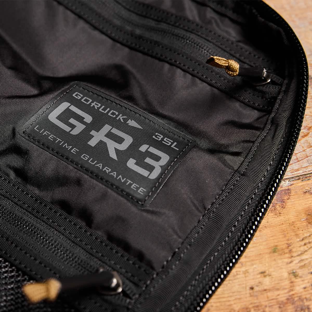 Close-up of a black GORUCK GR3 - Ripstop ROBIC® carry-on rucksack, displaying the label with GR3 35L and Scars Lifetime Guarantee. The top is partially open, showcasing a rugged zipper against a wooden surface.
