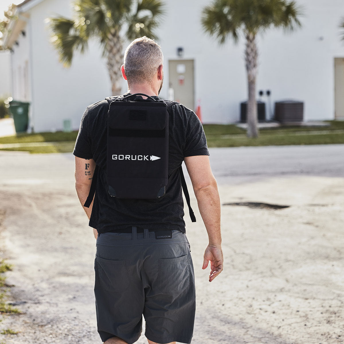 Man wearing a black Ruck Plate Carrier 3.0 made of durable ballistic nylon, walking on a sunny street with palm trees and buildings in the background.
