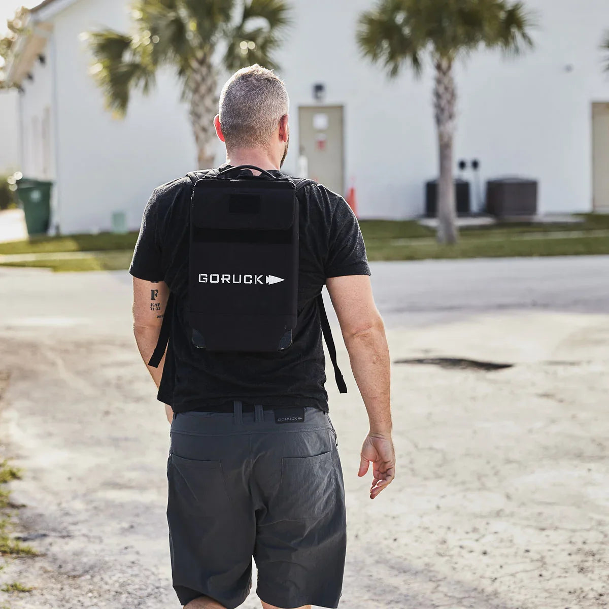 Man wearing black GORUCK backpack and gray shorts walking outdoors on a sunny day near palm trees