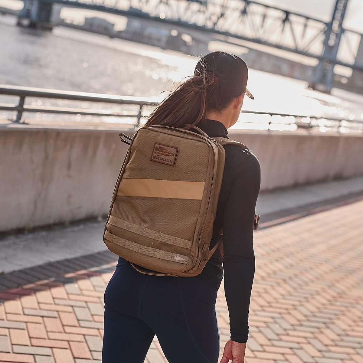 Person wearing a coyote brown GORUCK Rucker 4.0 15L backpack by a riverside walkway at sunset