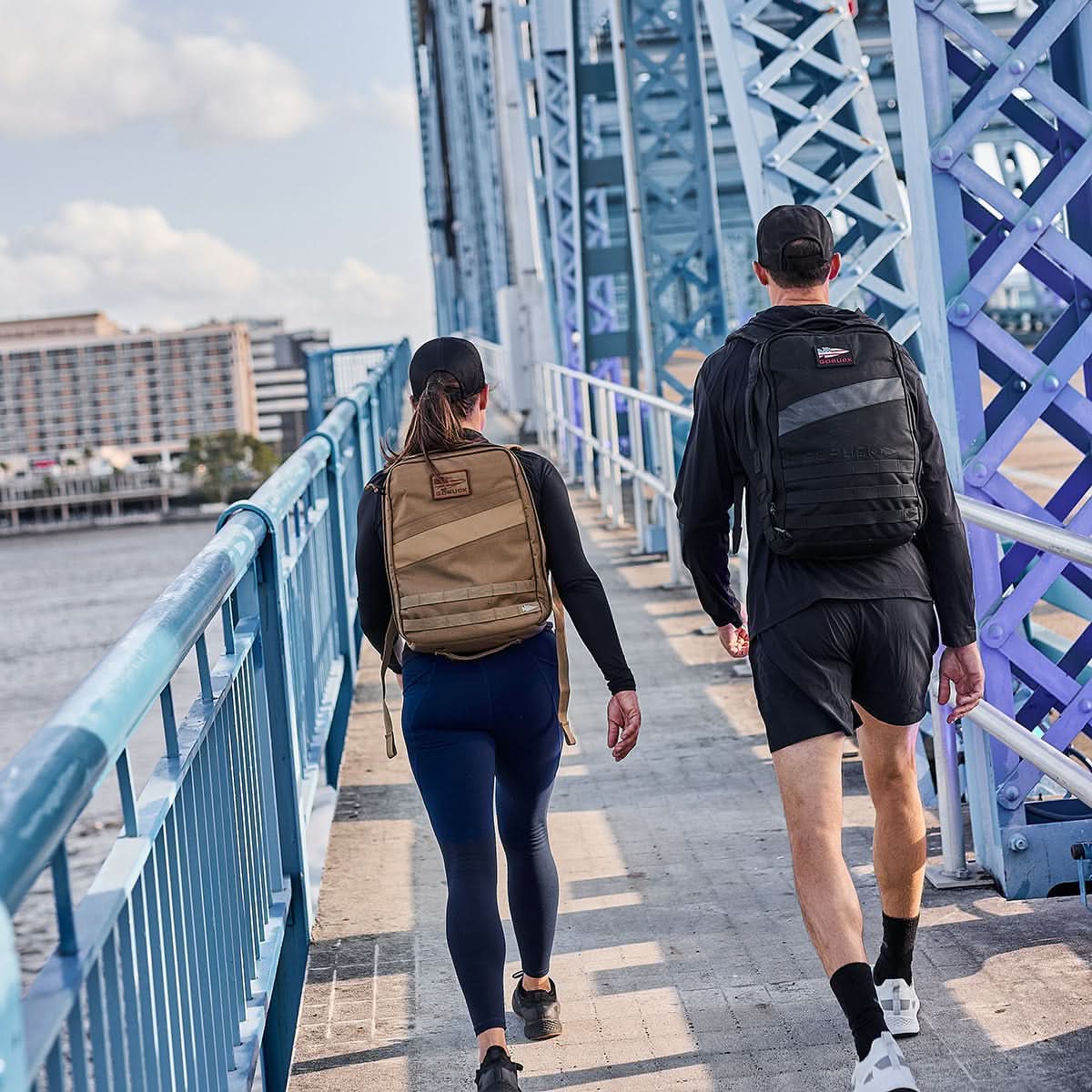 Two people carrying Rucker 4.0 backpacks ruck across a blue bridge on a sunny day, city buildings in the background—an ideal fitness activity.