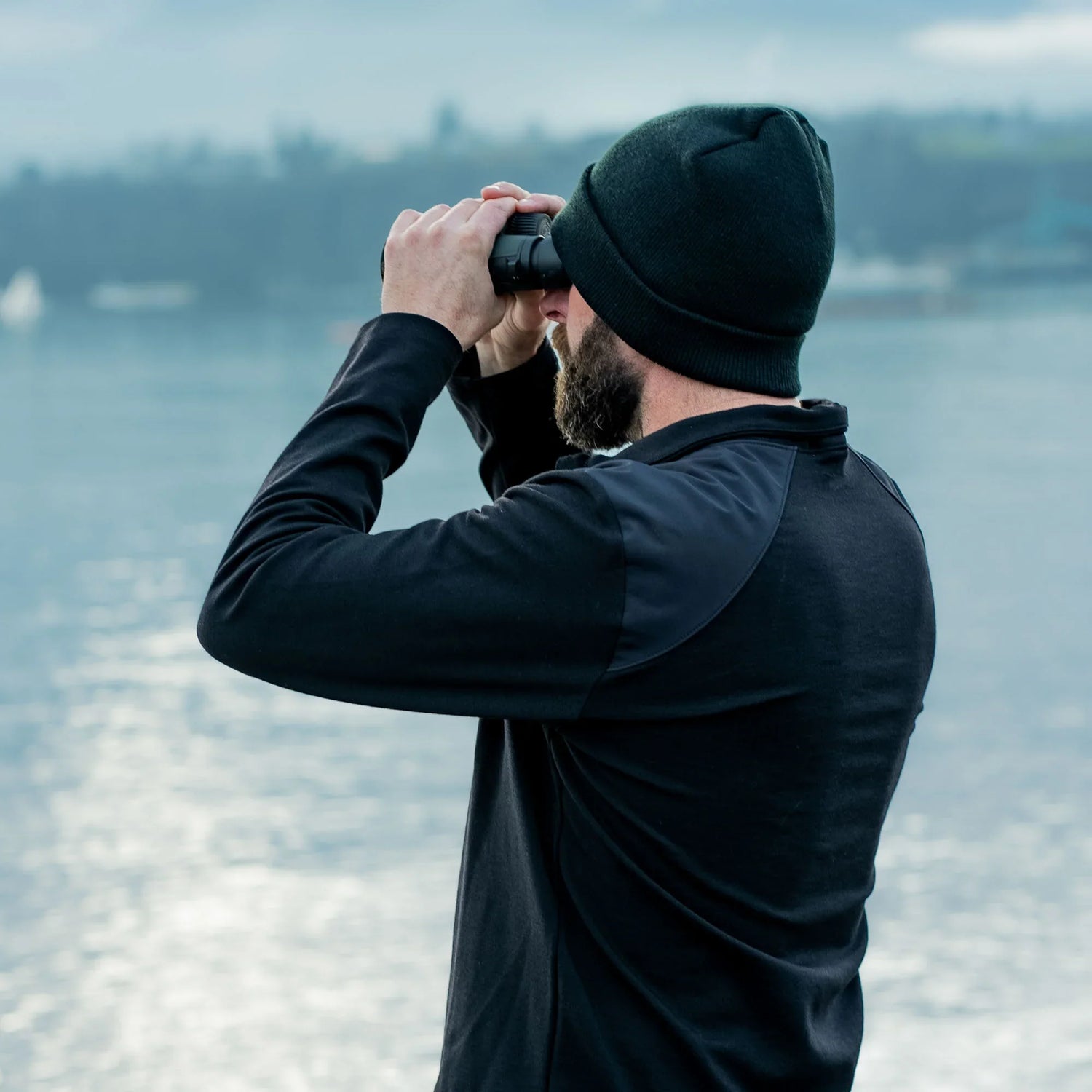 Man in GORUCK outdoor gear with beanie using binoculars by a lake
