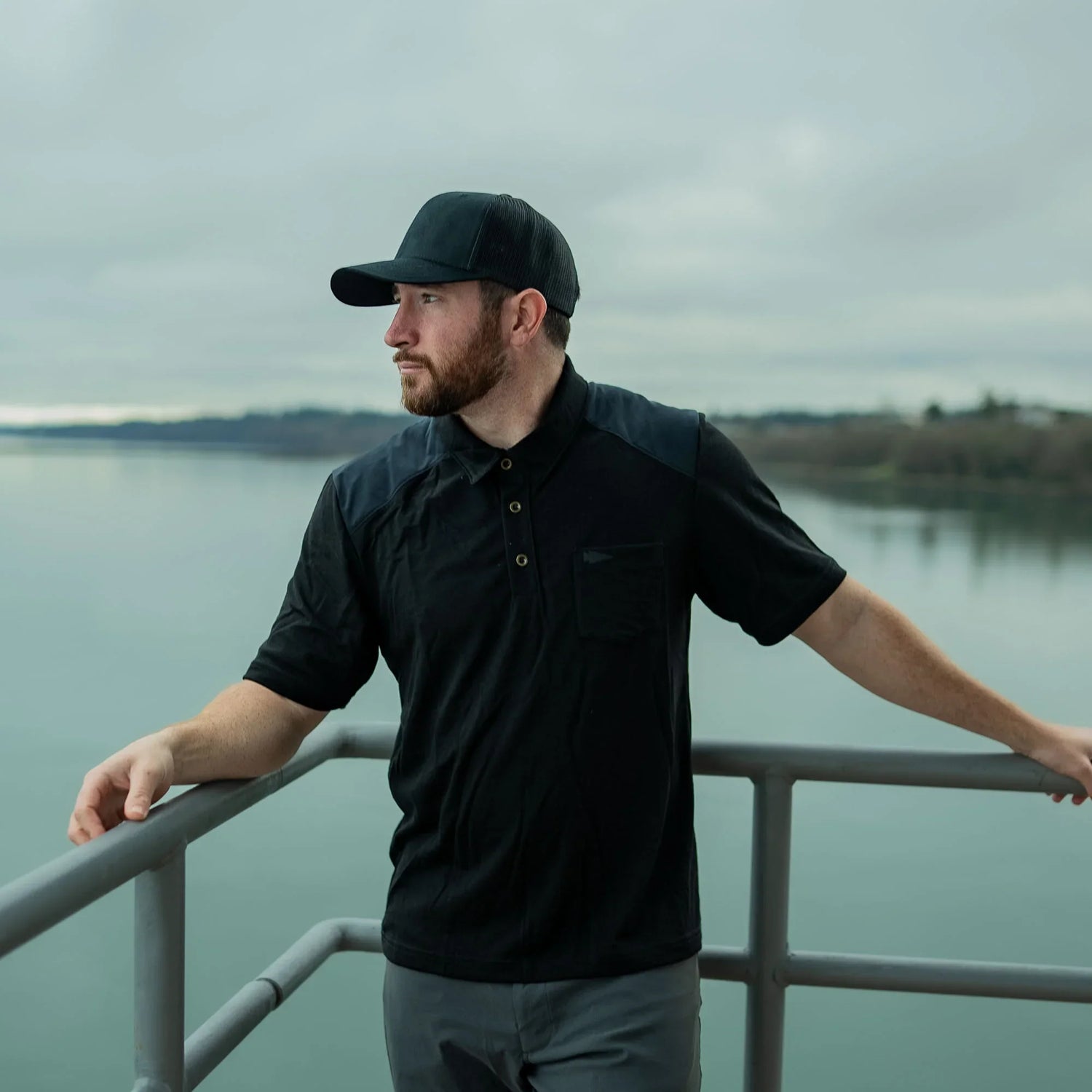 Man in GORUCK black polo and cap standing by water on an outdoor railing