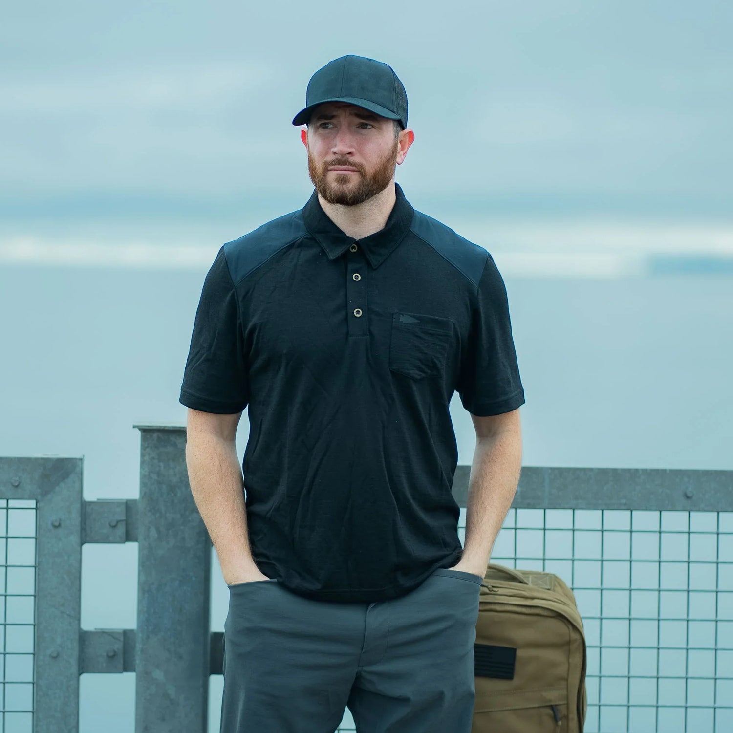 Man in black GORUCK polo and cap standing by railing outdoors, tan rucksack nearby