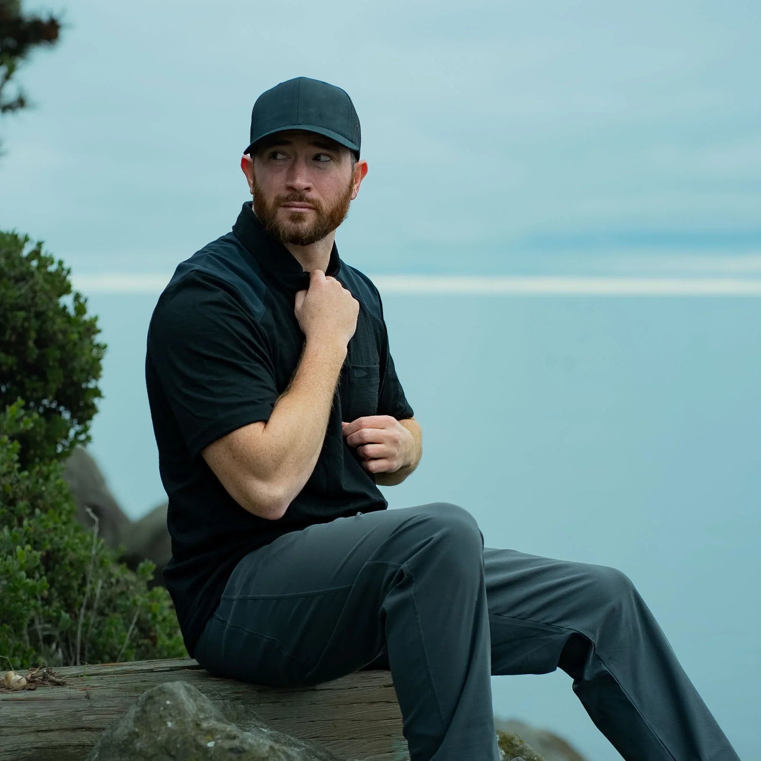 Man in GORUCK outdoor gear and black cap sitting on log by water