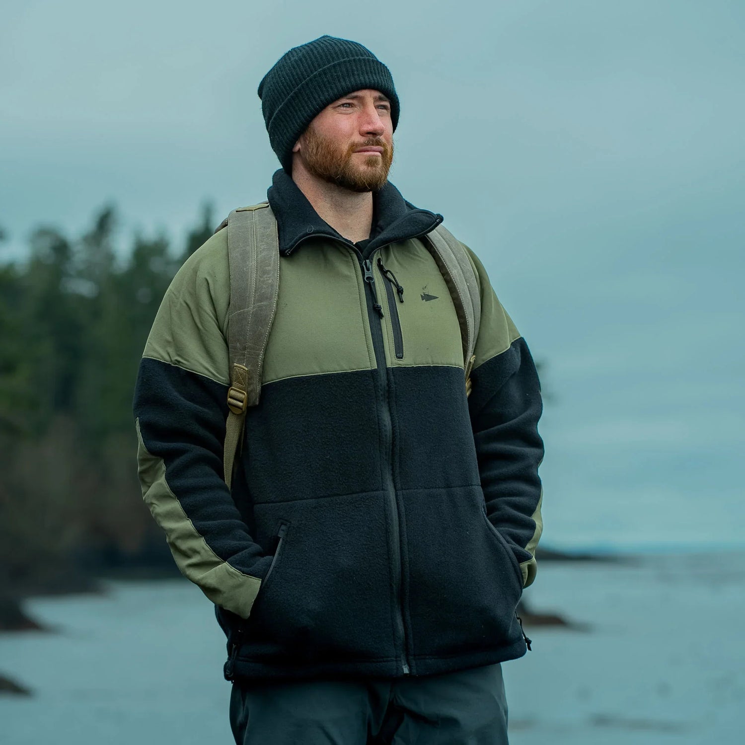 Man wearing GORUCK fleece jacket and backpack outdoors near water and forest