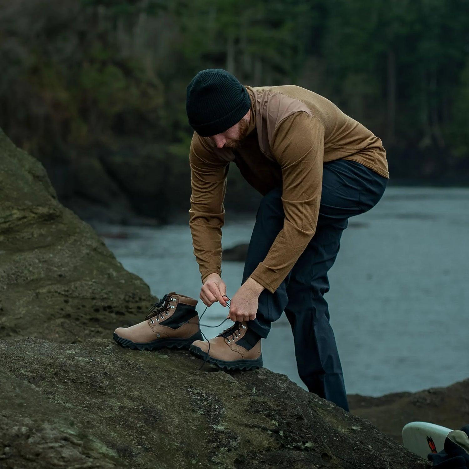 Man in outdoor gear tying durable rucking boots on rocky terrain near water