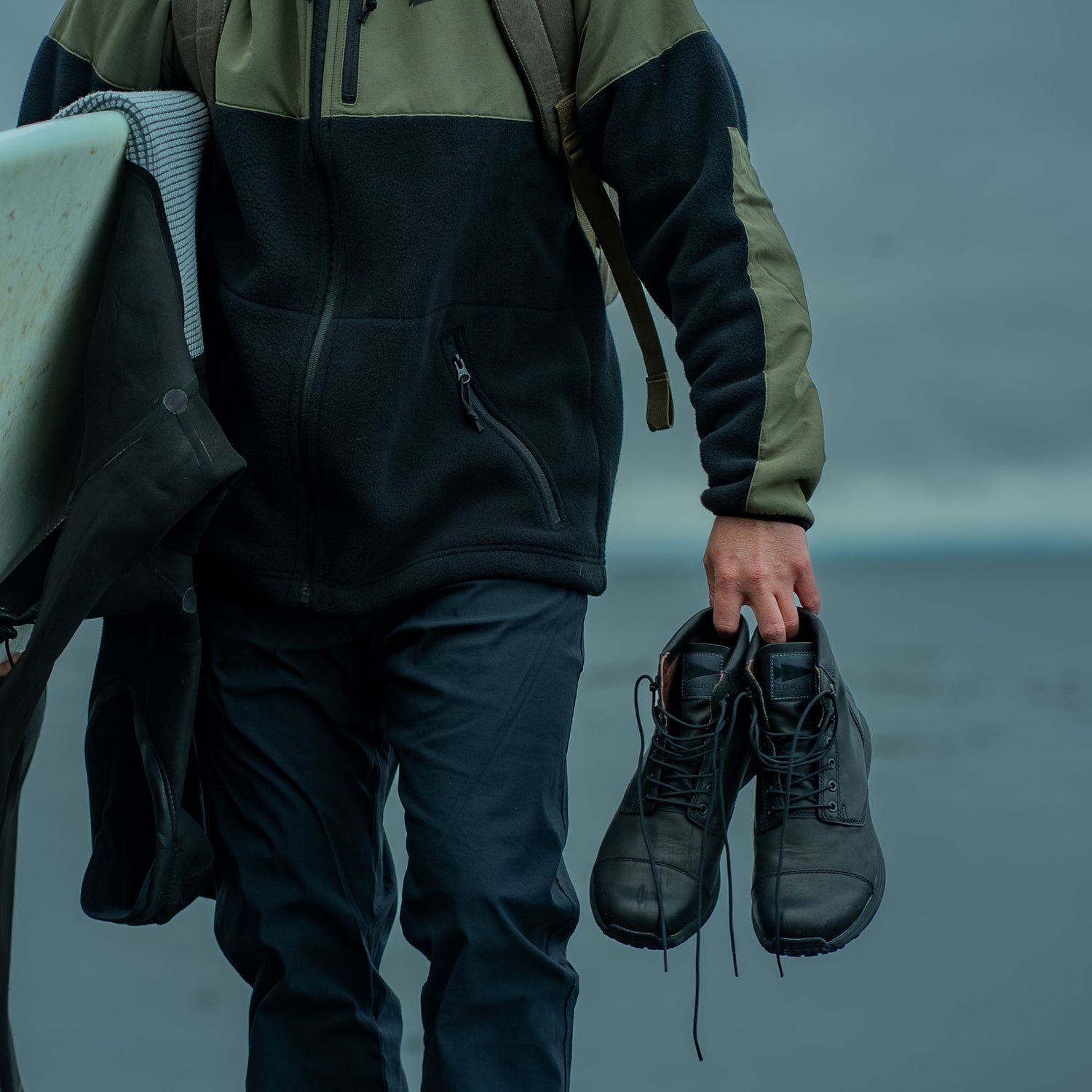 A person holding MACV-1 Traveler boots and a wetsuit, standing outdoors near water under a cloudy sky.