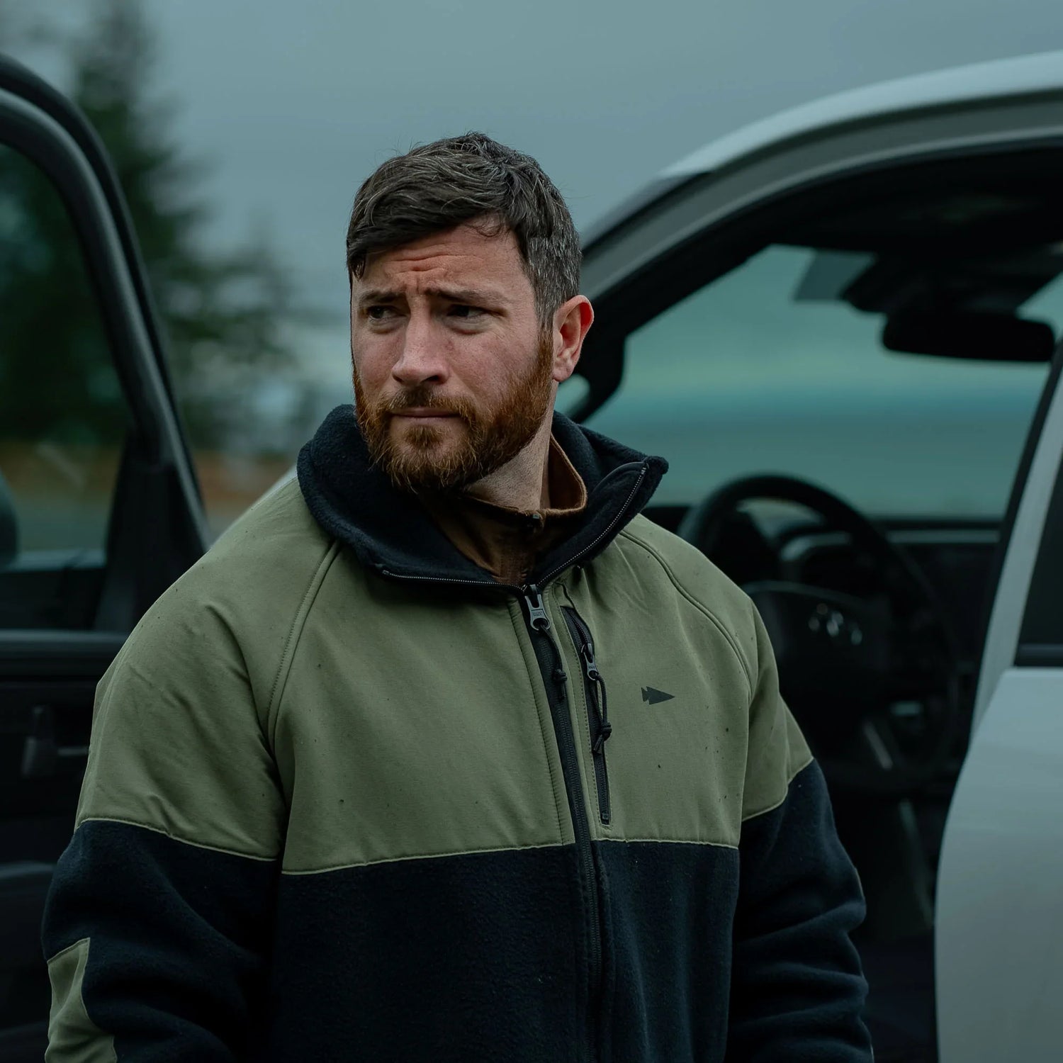 Man wearing green and black GORUCK jacket standing next to an open car door outdoors