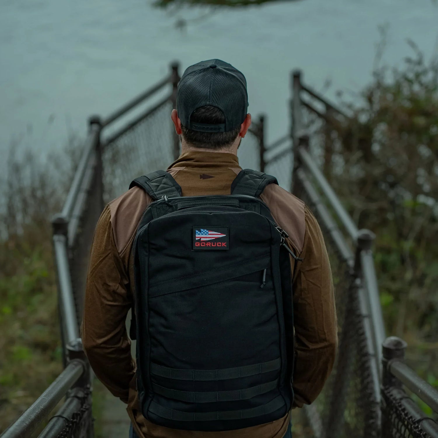 Man wearing a black GORUCK backpack and cap standing on a narrow metal bridge near water and foliage