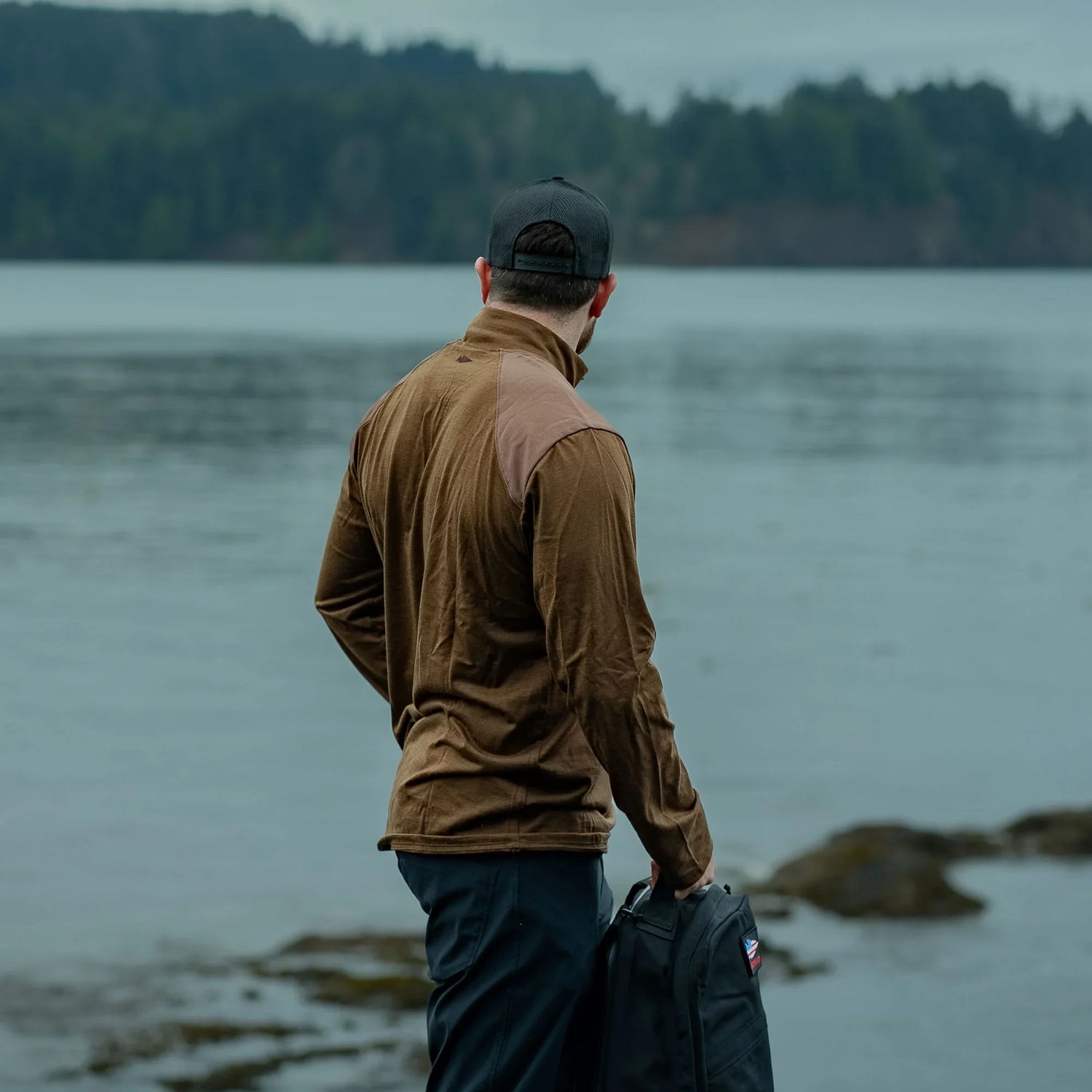 Man in brown GORUCK rucking jacket holding backpack by lake, outdoors, forest background