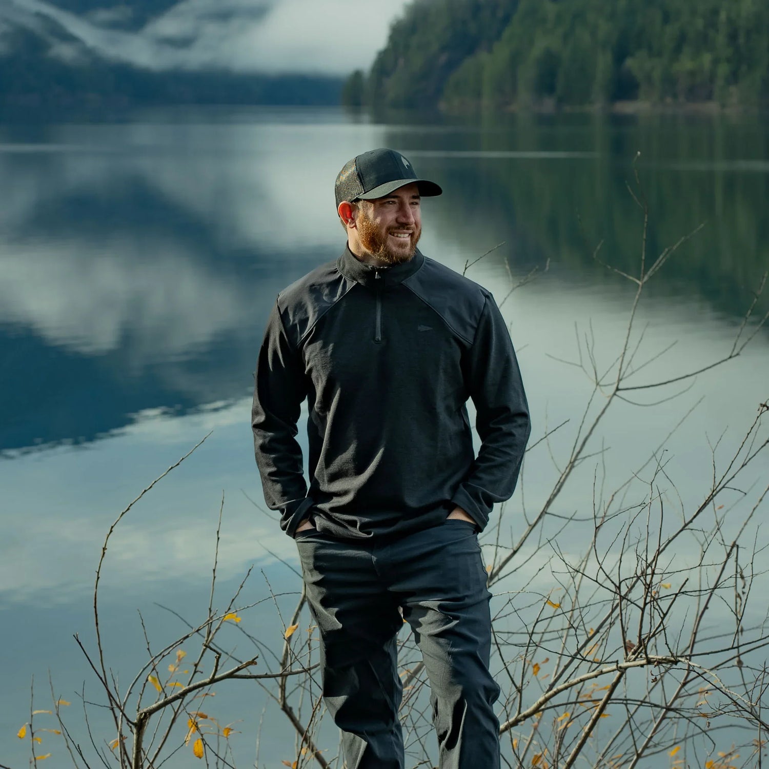 Man in black GORUCK outdoor gear smiling by a calm lake with pine forest backdrop