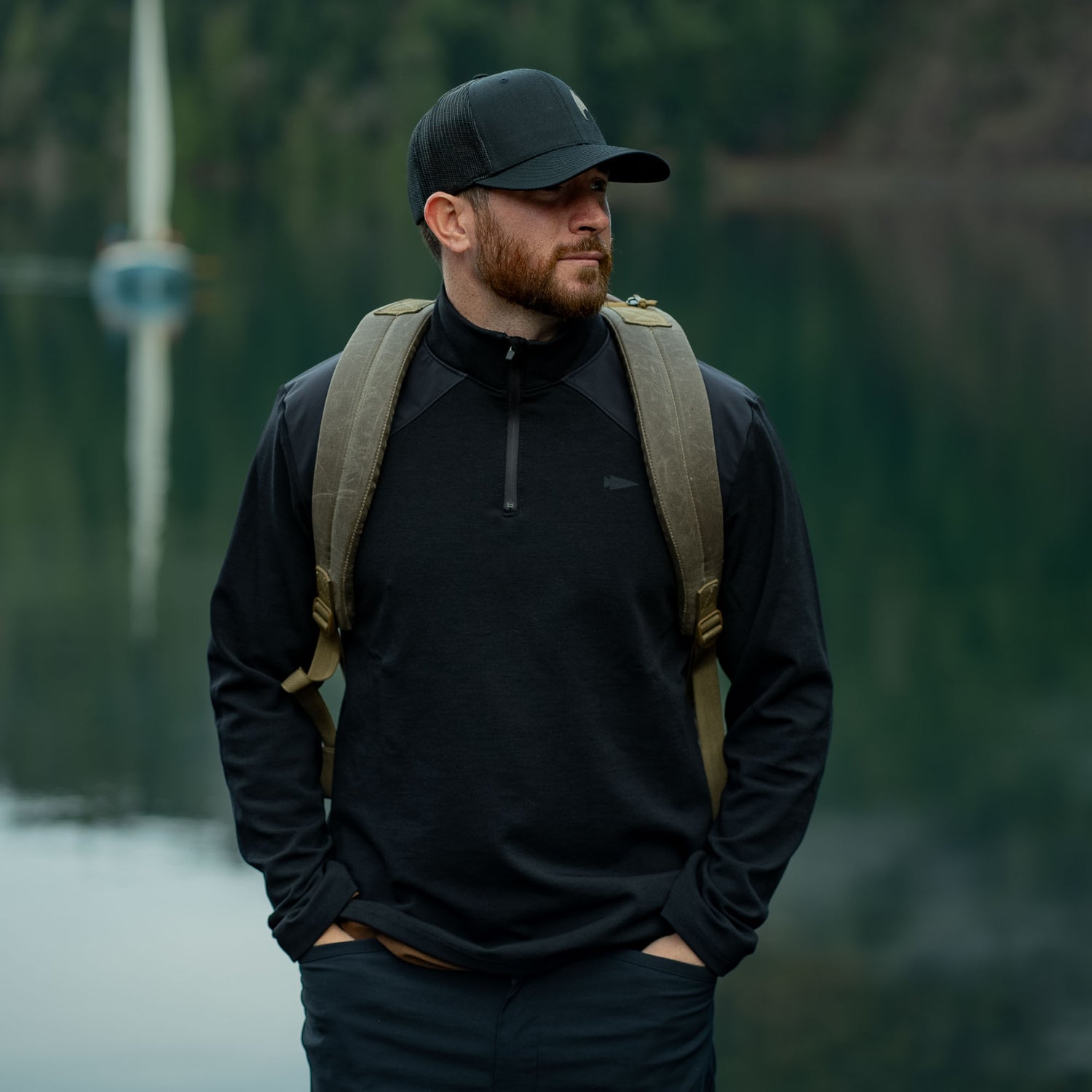 Man in GORUCK rucking gear with backpack, black pullover, and cap outdoors by a lake.