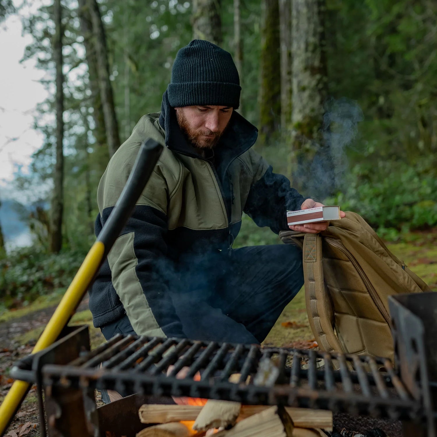 Man in outdoor gear by a campfire with GORUCK backpack in forest setting
