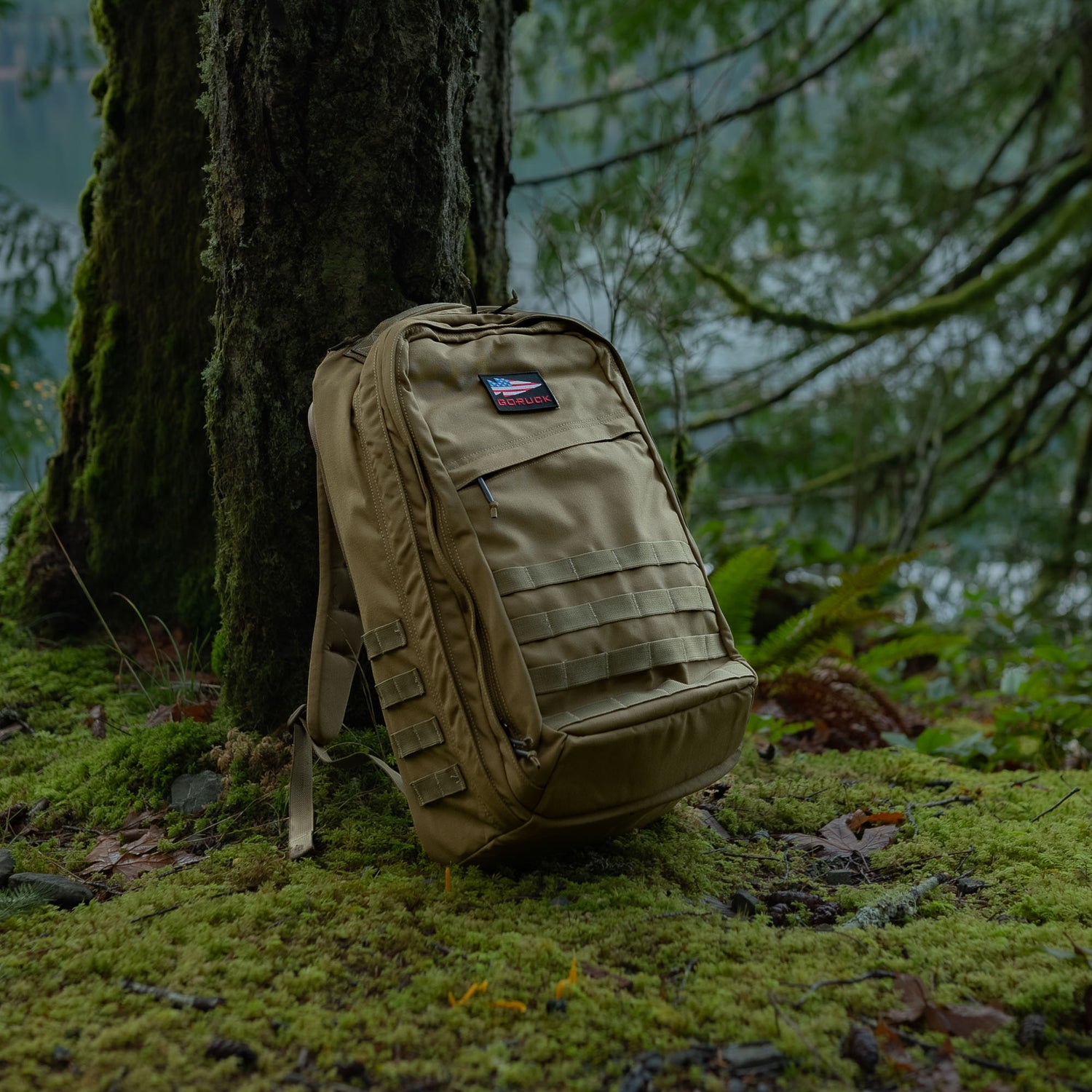 A GR2 USA - Cordura rucksack in tan rests against a mossy tree in a lush, green forest near a lake.