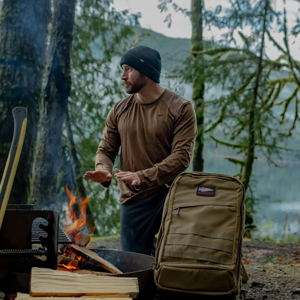 Man warming hands by campfire with GORUCK rucksack in forest near lake