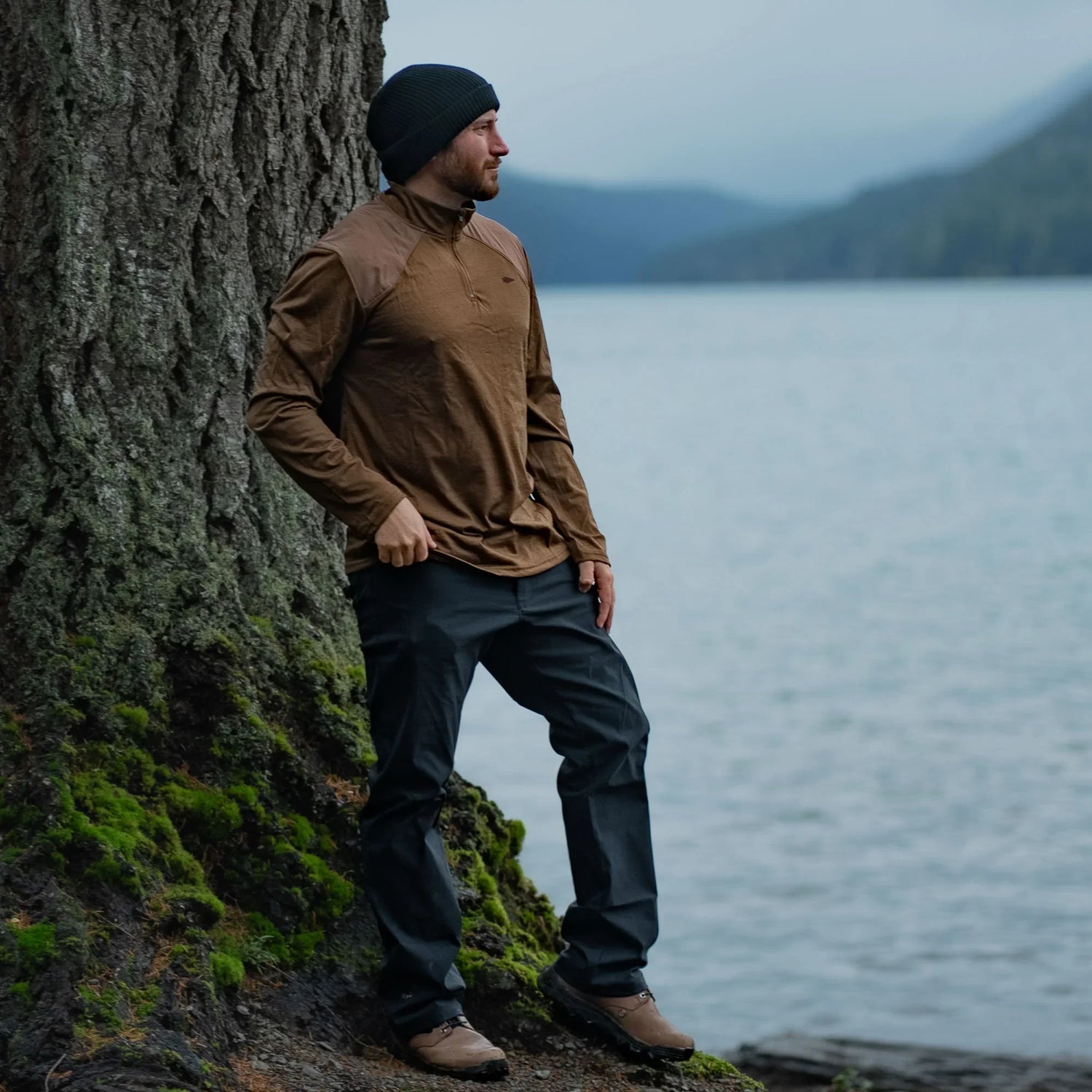 Man in GORUCK outdoor apparel standing by a tree beside a lake with mountains in the background