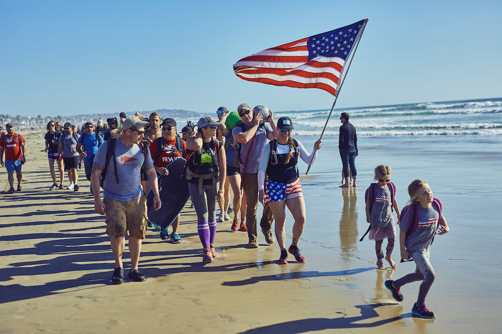 A group walks along a beach, one person holds a large American flag; children walk in front.