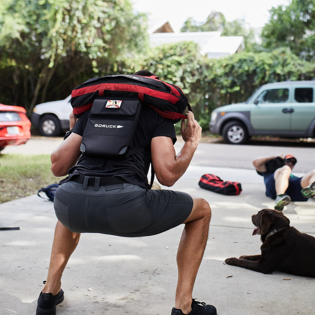 A person does squats outdoors with a weighted backpack in Men’s Simple Shorts - Lightweight ToughDry®, while a dog and another person rest nearby.