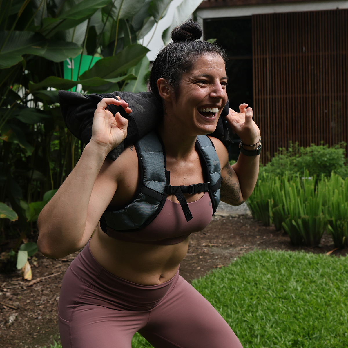 A woman in workout clothes smiles while squatting outdoors, showcasing the Spy Ruck weighted vest—a top choice in women’s fitness gear.