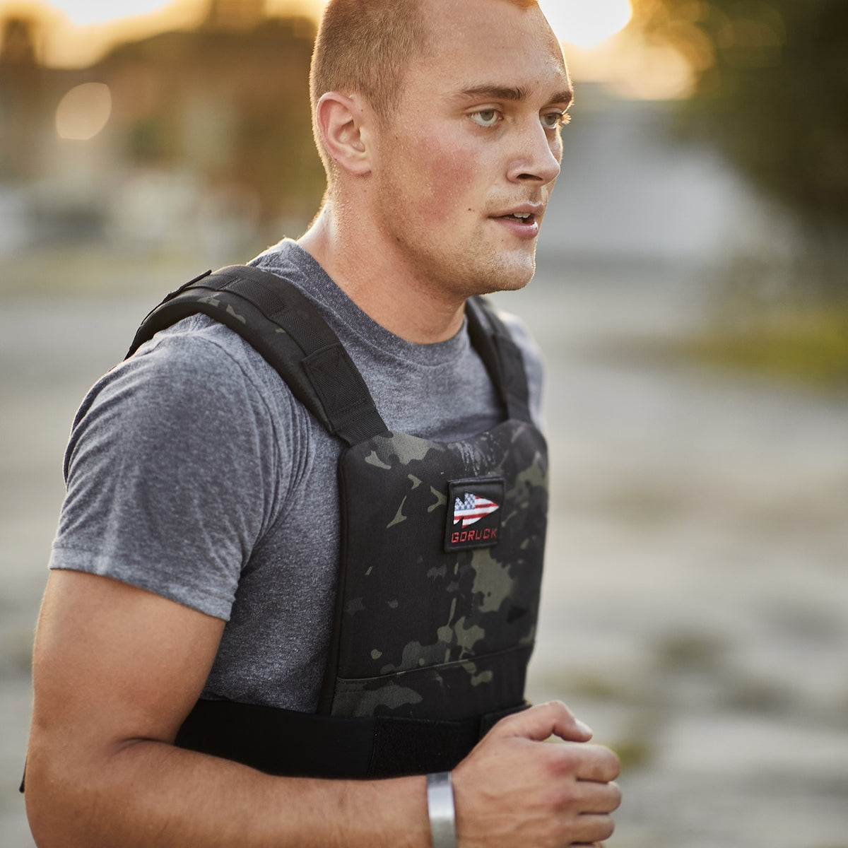A man in a grey t-shirt runs wearing a black camouflage GORUCK training weight vest with an American flag patch.