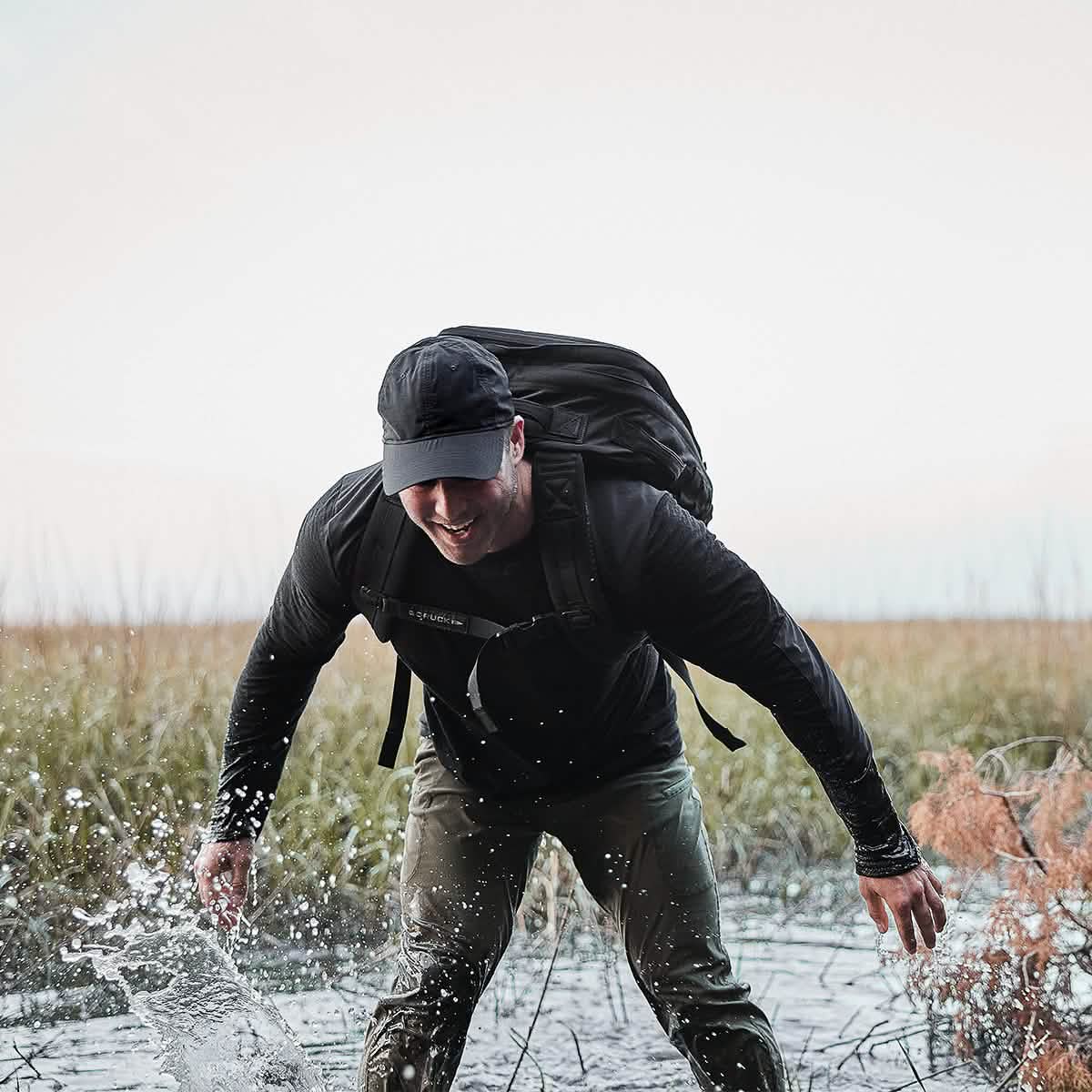 A person wearing a Performance TAC Hat - Slick from GORUCK and a shirt made of ToughDry® fabric playfully splashes water while walking through a shallow, grassy wetland. The sweat-wicking gear remains resilient under the overcast sky, with tall grass swaying in the background.