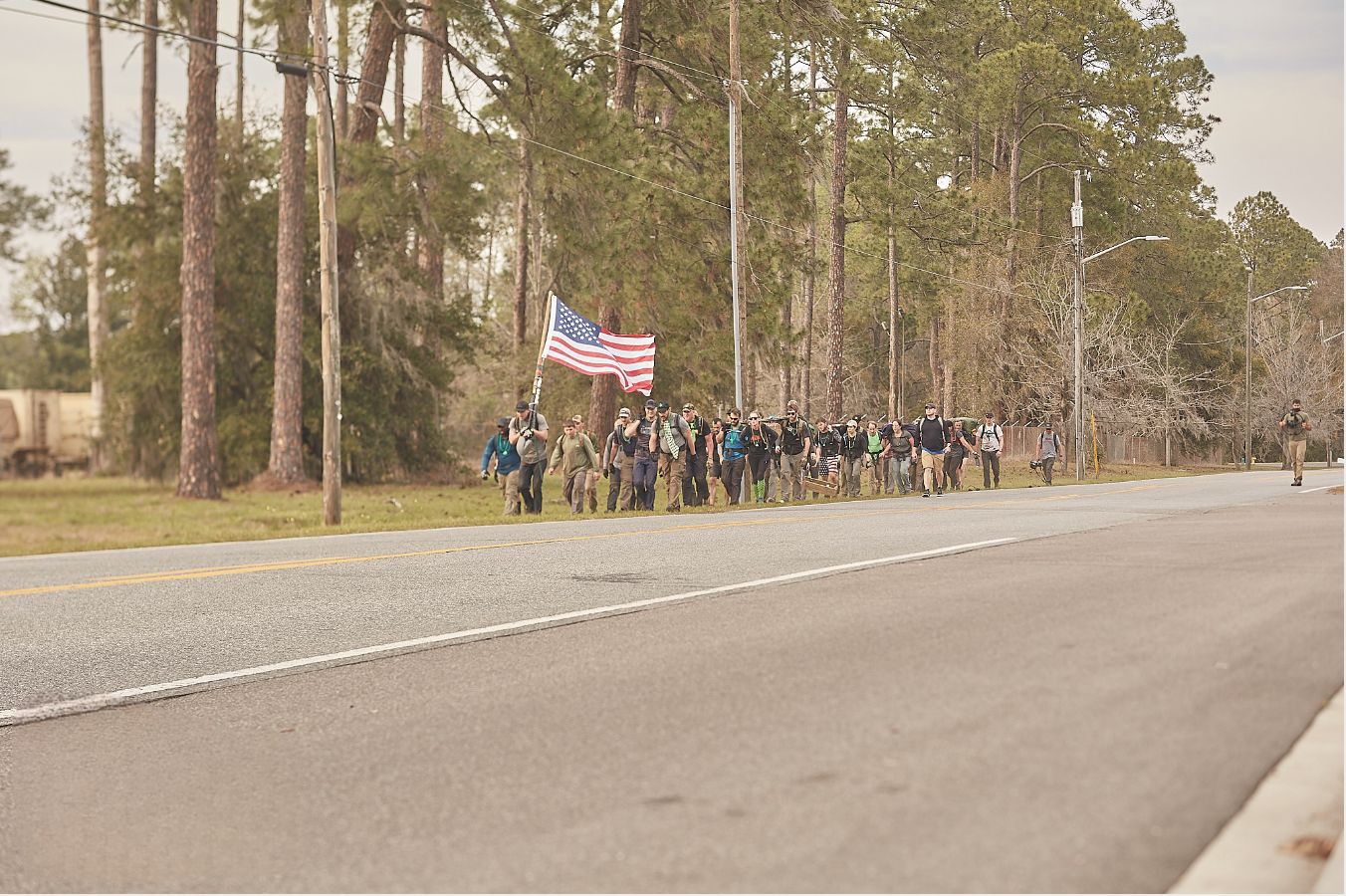 A group of people marches along a road, one carrying a large American flag, with trees in the background.