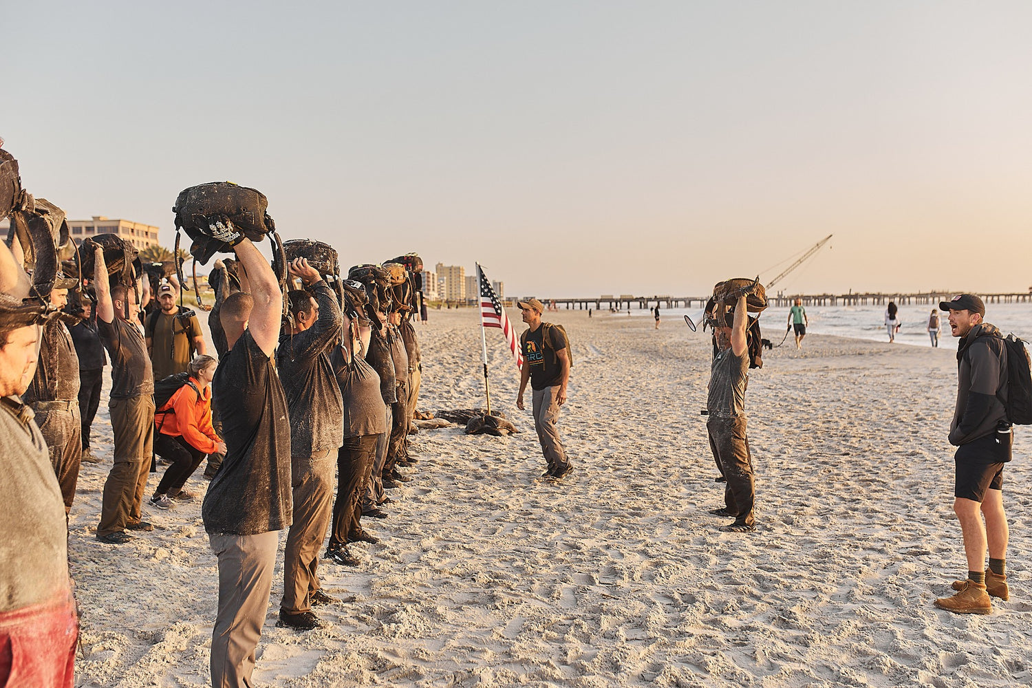 A group of people lift sandbags over their heads on a sandy beach during a fitness event at sunset.