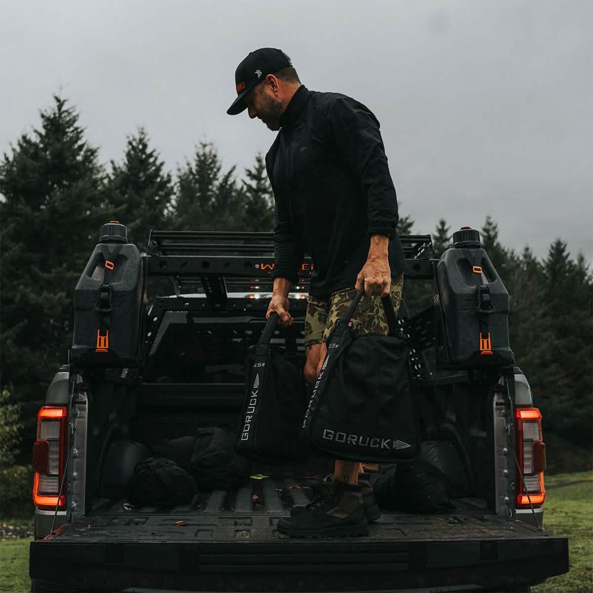 Man loading GORUCK black sand-filled Jerry cans into truck bed in outdoor forest setting