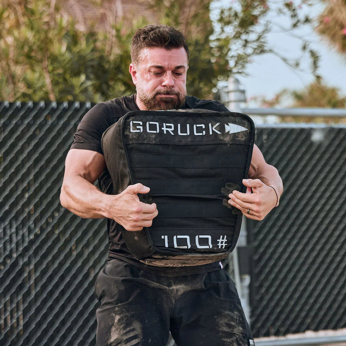 Man lifting dirty GORUCK weighted sandbag during outdoor rucking workout near fence
