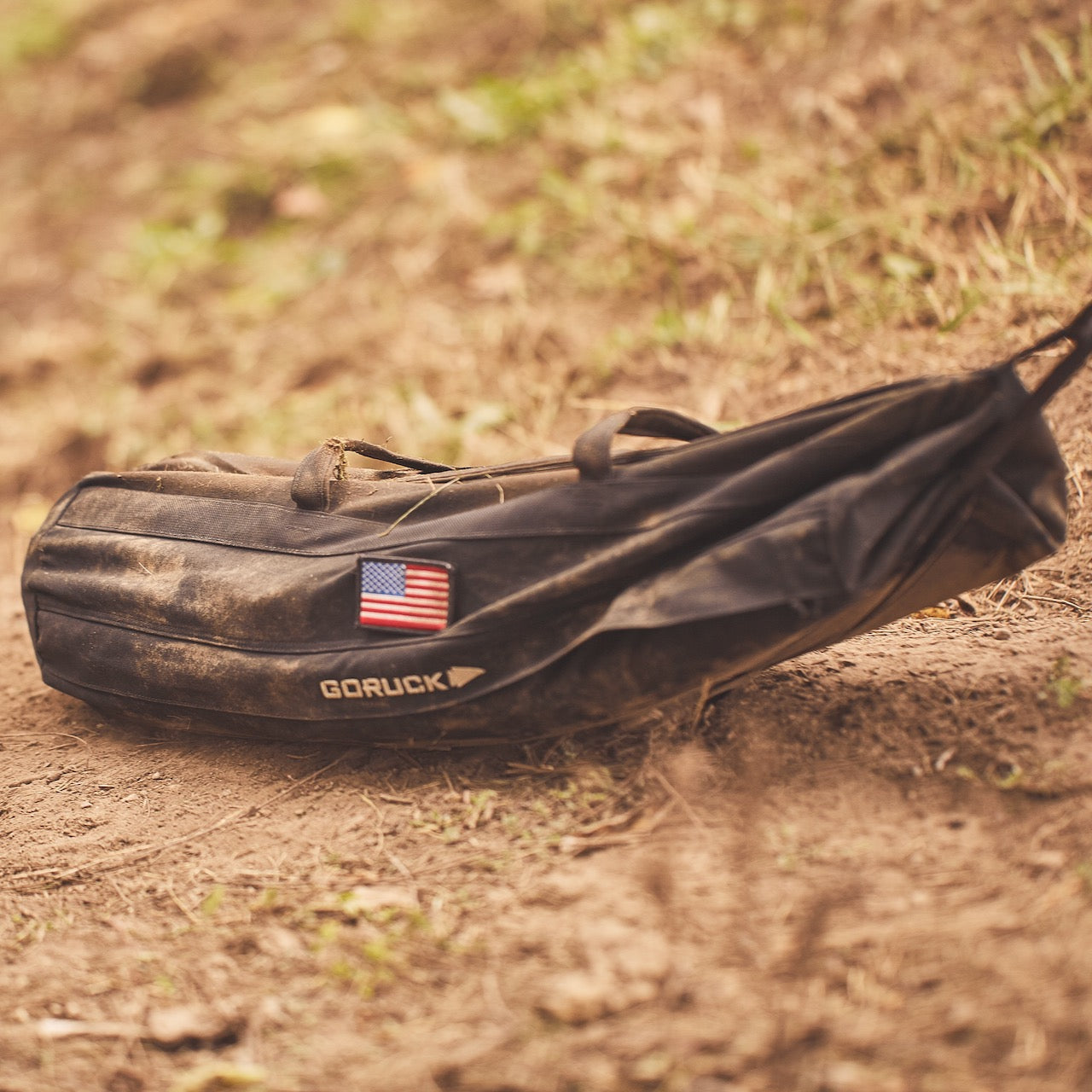 The Sandbag Kit (w/ Filler Bag), a black GORUCK bag featuring a U.S. flag patch and heavy-duty construction, rests on dirt next to some grass.