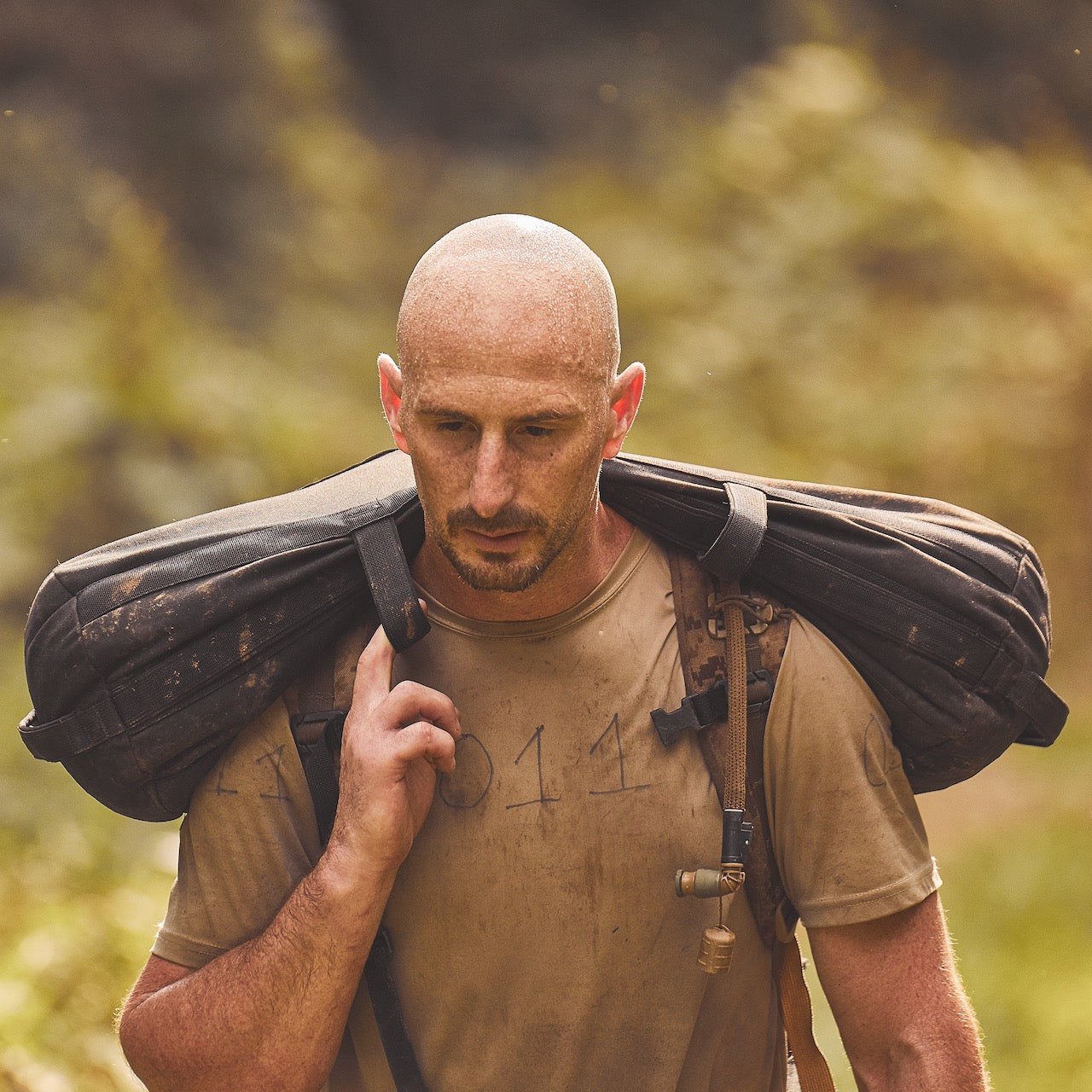 A bald man carries a Sandbag Kit (w/ Filler Bag) over his shoulders while walking outdoors in a sunlit, natural setting.