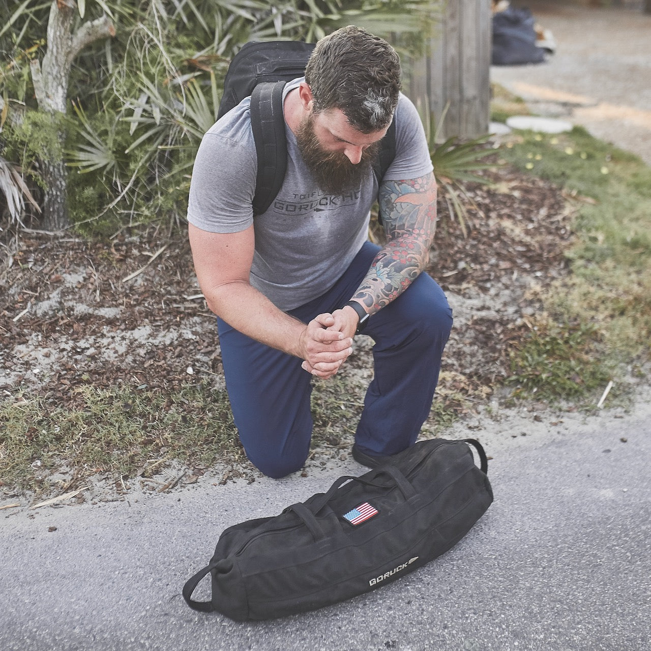 A man kneels outdoors, resting on a backpack with the Sandbag Kit (w/ Filler Bag) and a duffel bag in front of him—ideal for tough training sessions.