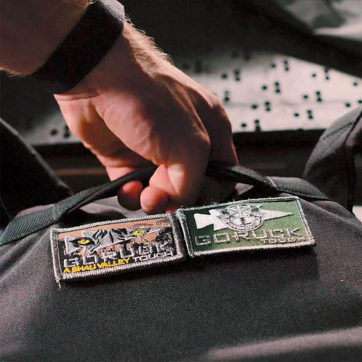 Close-up of a hand gripping a black GORUCK backpack with embroidered rucking patches