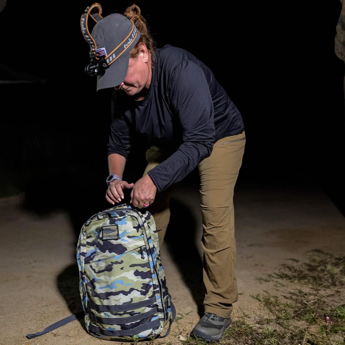 Person wearing headlamp and outdoor gear opening GORUCK camo backpack at night on dirt ground