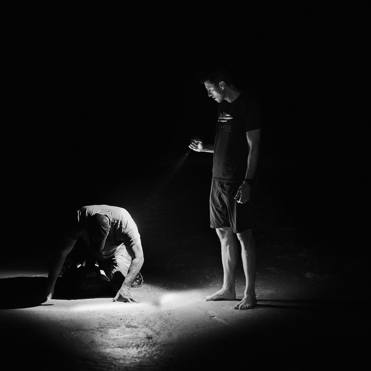 Two men in the dark; one stands with a flashlight, the other kneels in its beam, wearing Men’s Simple Shorts - Lightweight ToughDry® made from 2-way stretch fabric for comfort in challenging conditions.