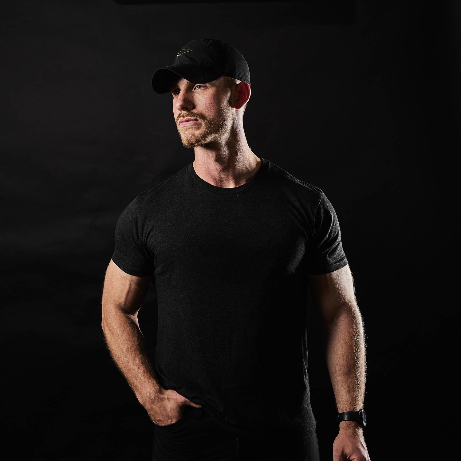 A man wearing the Slick Short Sleeve Tee - Tri-BlendX in black and a cap stands against a dark background, looking slightly to the side.