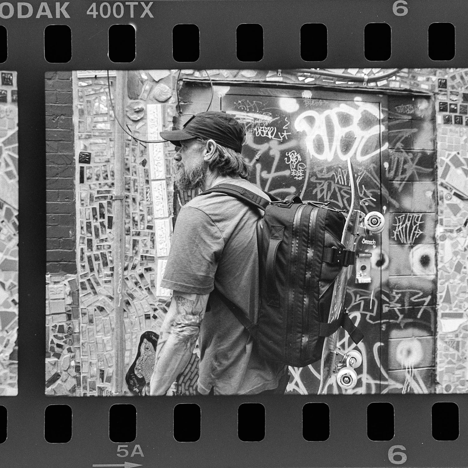 A man with a backpack stands by a graffiti wall in a black and white photo, highlighting his Skate Ruck—an urban skateboard backpack featuring a durable, bombproof laptop compartment.