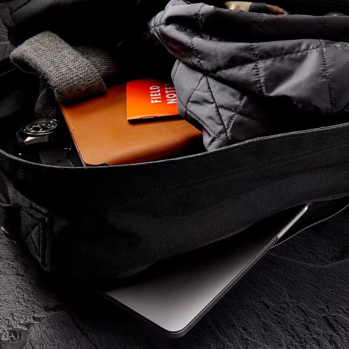 Open black rucking bag with gray jacket, watch, notebook, Field Notes notebook, and laptop inside
