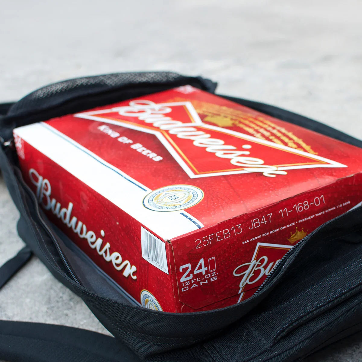 24-pack Budweiser beer can box partially inside an open black backpack on concrete