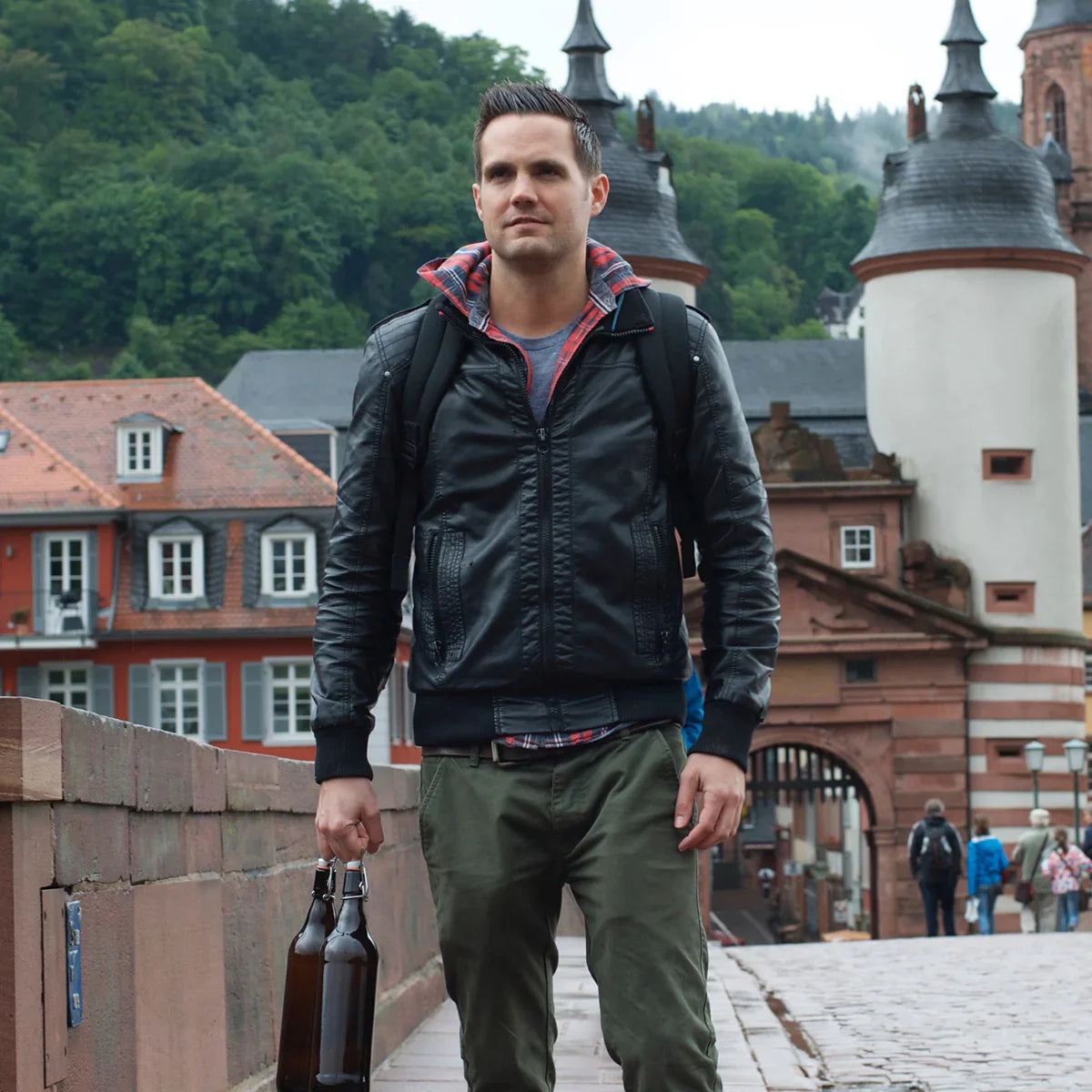 A man in a black jacket walks on a bridge, carrying bottles and the GR1 USA Slick - Ballistic Nylon Cordura backpack; historic buildings and trees create a scenic backdrop.