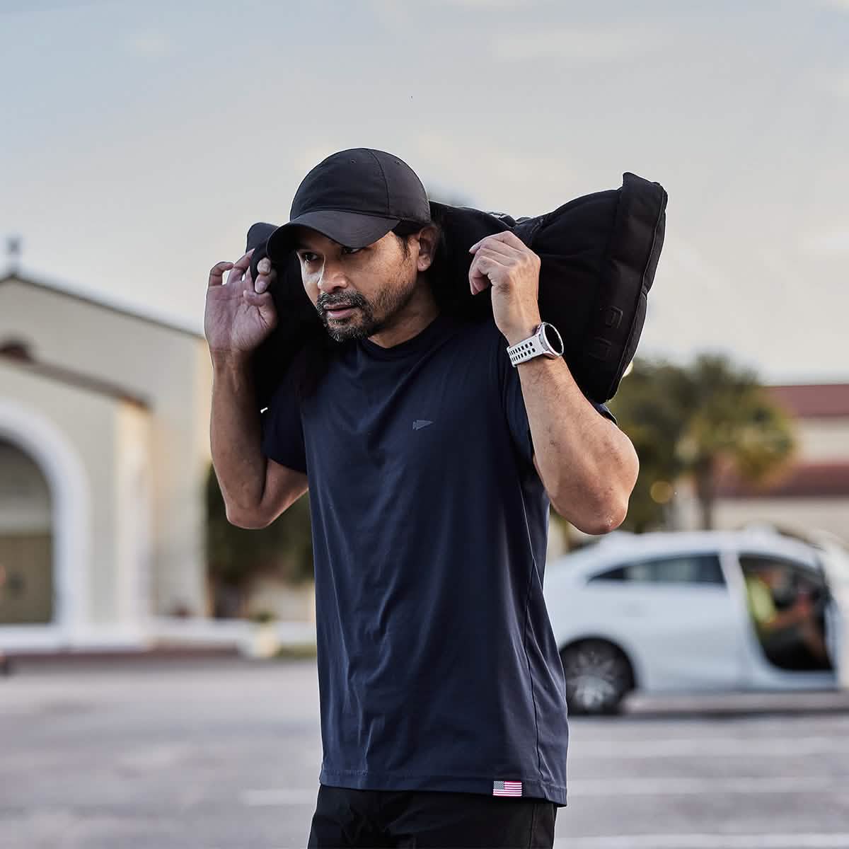 A man wearing a GORUCK Performance TAC Hat - Slick with TOUGHDRY technology and a navy blue t-shirt carries a heavy black sandbag across his shoulders. His sweat-wicking gear keeps him comfortable outdoors, where a blurred building and white car are in the background. A smartwatch adorns his left wrist.