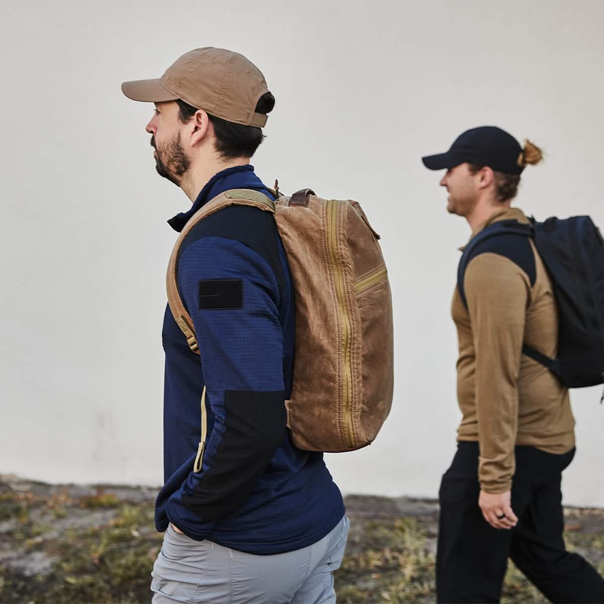 Two men are walking side by side, each wearing a GORUCK Performance TAC Hat - Slick - TOUGHDRY and carrying backpacks. The man in the foreground sports a beige backpack and a blue shirt, while the man behind carries a black backpack and wears a tan sweater. Their gear is ideal for outdoor adventures with its sweat-wicking ToughDry® fabric.