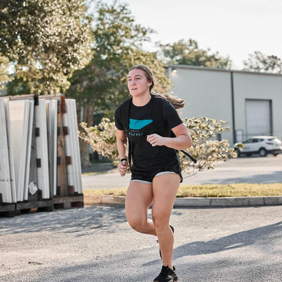 Athletic woman running outdoors wearing black GORUCK Spearhead T-shirt and black shorts with a backpack