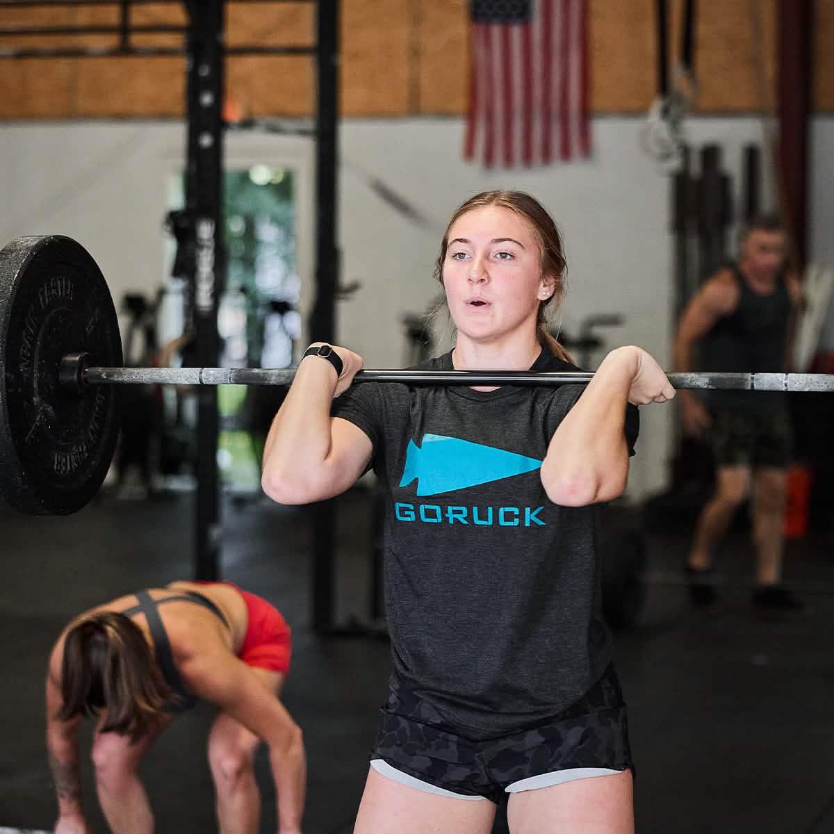Young woman lifting barbell in gym wearing black GORUCK t-shirt with blue logo, American flag in background