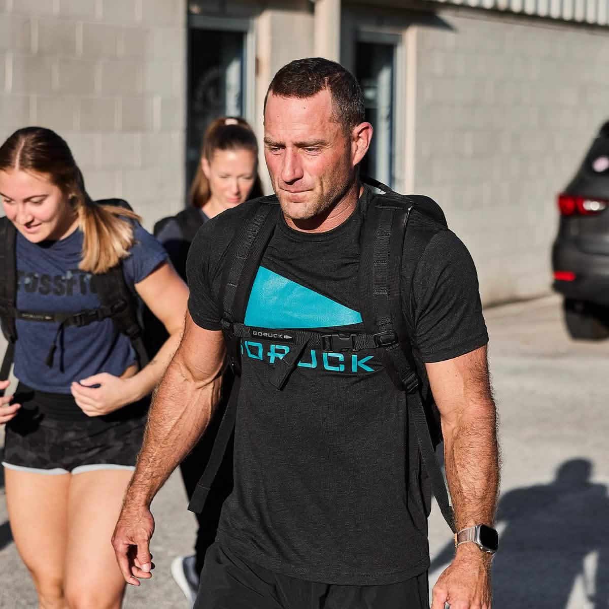 Man wearing black GORUCK t-shirt with blue logo and backpack outdoors, focused during group fitness activity